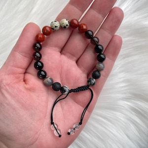 Puede incluir: Un brazalete de cuentas con piedras negras, marrones, rojas y blancas. El brazalete tiene un cordón negro con cuentas transparentes en los extremos.