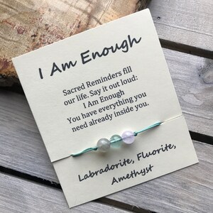 May include: A light blue string bracelet with three gemstone beads: labradorite, fluorite, and amethyst. The bracelet comes with a card that reads "I Am Enough" and includes a message about sacred reminders.