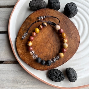 Puede incluir: Un brazalete de piedras preciosas multicolores con cuentas marrones, amarillas y rojas. El brazalete también incluye cuentas de cuarzo transparente y de roca volcánica negra.