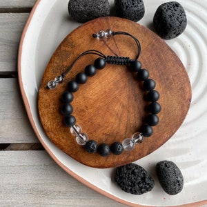 May include: A black and clear beaded bracelet with a macrame knot closure. The bracelet is made with black onyx, clear quartz, and lava rock beads. The bracelet is displayed on a wooden surface.