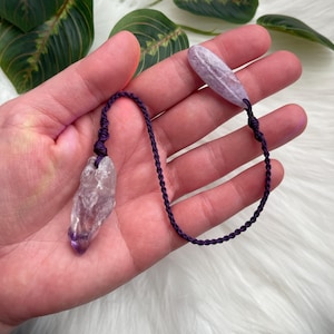 May include: A purple amethyst crystal pendant with a dark purple cord. The crystal is a light purple color with a darker purple tip. The cord is knotted at both ends, with a smaller crystal at the other end.