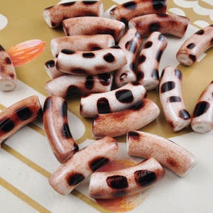 Puede incluir: Una colección de cuentas de cerámica marrón y blanca con un patrón de estampado de leopardo. Las cuentas tienen forma de cilindro curvo y miden aproximadamente 2,5 cm de largo.