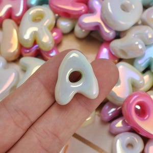 May include: A collection of iridescent alphabet beads in various pastel colours including pink, white, and light blue. Each bead features a rounded, puffy letter shape with a central hole for threading, showcasing a glossy, pearlescent finish.