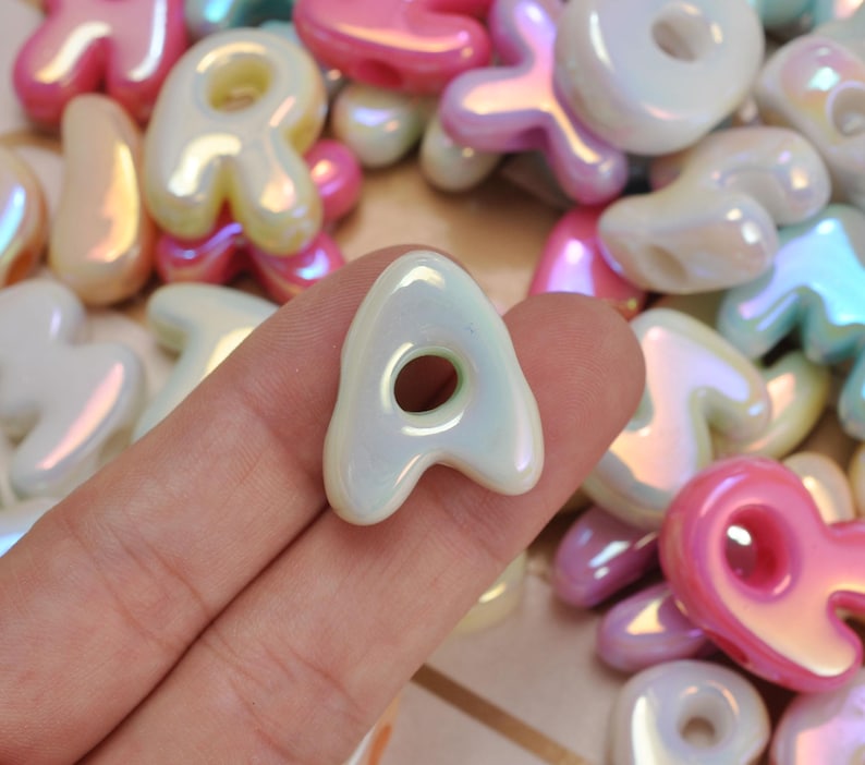 May include: A collection of iridescent alphabet beads in various pastel colours including pink, white, and light blue. Each bead features a rounded, puffy letter shape with a central hole for threading, showcasing a glossy, pearlescent finish.