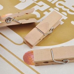 Puede incluir: Tres pinzas de madera con clips de resorte de metal. Las pinzas son de color madera natural y están dispuestas sobre una superficie rayada.