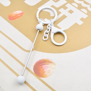 Pode incluir: Chaveiro de metal branco com uma haste longa, fina e prateada e duas contas redondas brancas. O chaveiro tem um anel de metal branco e um gancho de metal branco.