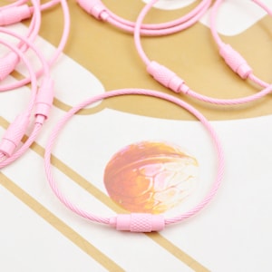 Peut inclure: Gros plan sur plusieurs porte-clés en métal rose avec un design de câble tressé. Les porte-clés sont posés sur une surface rayée.