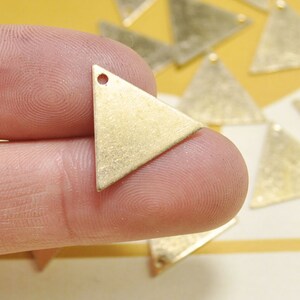May include: A close-up of a gold triangle pendant with a small hole at the top. The pendant is made of metal and has a textured surface.