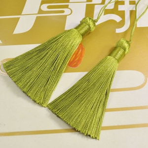 May include: Two green tassel ornaments with a long, thin, fringed design. The tassels are made of a soft, silky material and are tied with a thin string.
