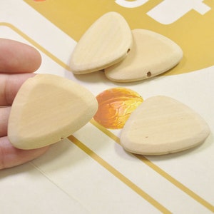 Puede incluir: Cuatro púas de guitarra de madera sin terminar en forma de triángulo. Las púas son de color marrón claro y tienen un agujero en el centro.