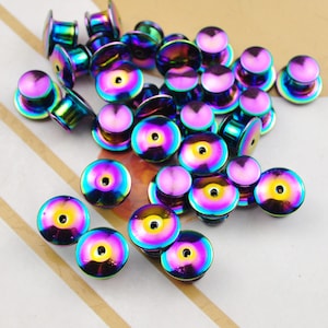 May include: A pile of iridescent rainbow colored metal safety pin backs. The pin backs are round and have a small hole in the center.