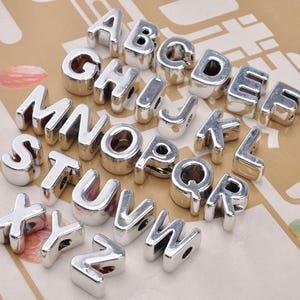 Op de afbeelding: Een verzameling van zesentwintig individuele zilverkleurige alfabetletterkralen, elk met een gladde, reflecterende metallic afwerking en een gat om te rijgen. De letters A tot en met Z zijn in een reeks gerangschikt en tonen een afgeronde, driedimensionale blokletterstijl.