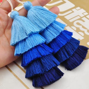 May include: Two blue and light blue ombre tassel trims with multiple layers. The tassels are made of soft, fluffy yarn and are perfect for adding a touch of colour and texture to any project.