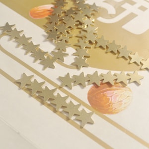 May include: A close-up of a white surface with a scattering of gold star-shaped embellishments. The stars are arranged in a line and a cluster.
