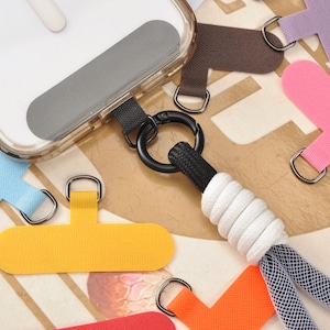 以下が含まれることがあります： グレーの粘着パッドが付いた透明な携帯電話ケースが、黒いカラビナと白と青のストラップで表示されています。 青、黄、ピンク、オレンジ、緑、紫のカラフルなT字型のタブが散らばっています。