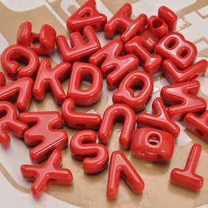 May include: A collection of glossy red plastic alphabet beads, each featuring a central hole for threading. The assortment includes various uppercase letters scattered in a pile, showcasing a smooth, reflective surface and rounded edges.