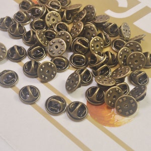 May include: A large quantity of antique gold-toned metal pin backs. The pin backs have a round shape with a small hole in the center and a raised, textured design around the edge.