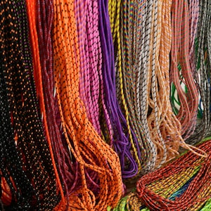 May include: A collection of multi-coloured elastic cords hanging vertically in a row. The cords feature various patterns including solid colours, speckled designs, and striped textures in shades of black, orange, purple, yellow, pink, blue, and green.
