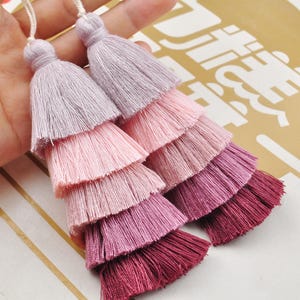 May include: Two multi-colored tassel ornaments with five layers of fringe. The tassels are a gradient of pink, purple, and burgundy.