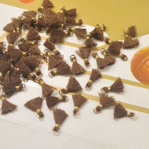 May include: A collection of brown tassel charms with gold accents. The tassels are scattered on a white surface.