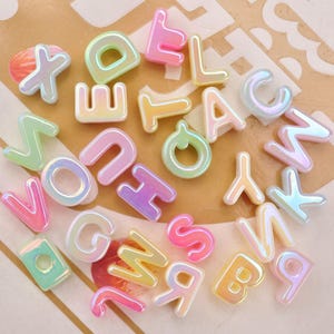 Op de afbeelding: Een verzameling individuele alfabetletters gemaakt van glad, iriserend plastic in diverse pastelkleuren zoals roze, geel, groen en blauw. Elke letter heeft een afgeronde, bolle vorm met een glanzende afwerking die het licht reflecteert.