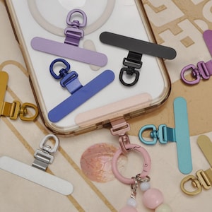 May include: A selection of colourful phone charms in shades of gold, silver, black, blue, and pink. Each charm has a rectangular base and a metal loop for attaching to a phone case or accessory. These charms are designed to be both stylish and functional.
