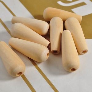 Könnte beinhalten: Eine Sammlung unfertiger Holzknöpfe, jeder mit einem abgerundeten Oberteil und einem sich verjüngenden Körper. Die Knöpfe haben eine helle, natürliche Holzfarbe und ein kleines Loch in der Unterseite. Sie sind auf einer weiß-gold gestreiften Oberfläche angeordnet.