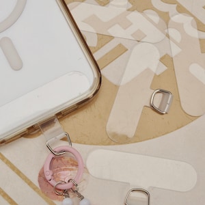 Op de afbeelding: Transparante telefoonhoes met een goudkleurige rand, een roze karabijnhaak en transparante plakstrips. De karabijnhaak heeft een kleine bedel met witte kralen. De hoes is ontworpen om een telefoon te bevatten.