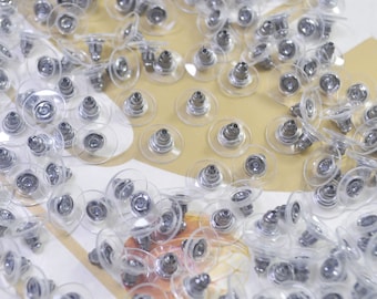 May include: A close-up of a large quantity of clear plastic earring backs with silver metal posts. The backs are scattered on a white surface.