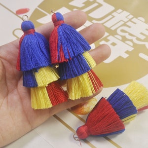 May include: Three layered tassels in red, yellow, and blue colors. The tassels are made of soft yarn and have a gold loop at the top.