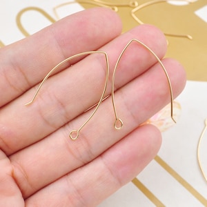 Peut inclure: Gros plan sur des crochets de boucles d'oreilles dorés. Les crochets ont une forme incurvée en V avec une petite boucle en bas pour attacher des perles ou des breloques. Les crochets sont tenus dans une main, suggérant leur taille et leur utilisation en bijouterie.