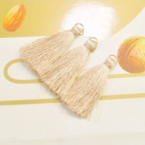 Può includere: Tre nappine beige con anelli in metallo dorato. Le nappine sono fatte di fili morbidi e setosi e sono lunghe circa 5 cm.