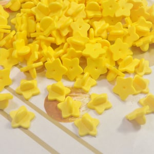 May include: A pile of bright yellow star-shaped pin backs. The pin backs feature a five-point star design with a small post and a clasp on the reverse. The objects are scattered on a light-coloured surface.