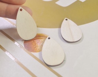 May include: Three unfinished wooden teardrop-shaped pendants with a hole at the top. The pendants are on a white and brown striped surface.