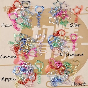 Könnte beinhalten: Eine Sammlung bunter Metall-Schlüsselanhänger in verschiedenen Formen, darunter Bären, Sterne, Kronen, D-Formen, Äpfel und Herzen. Die Schlüsselanhänger sind in Rot-, Rosa-, Blau-, Grün- und Goldtönen gehalten.