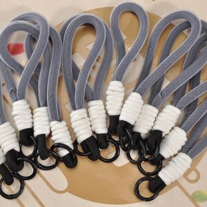 May include: A collection of gray and white keychains. Each keychain features a gray woven loop, a white wrapped section, a black cord, and a black metal ring. The keychains are arranged on a light-colored surface.