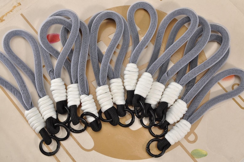 May include: A collection of gray and white keychains. Each keychain features a gray woven loop, a white wrapped section, a black cord, and a black metal ring. The keychains are arranged on a light-colored surface.