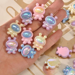 Puede incluir: Una colección de adornos iridiscentes con forma de alienígena en colores pastel. Las pequeñas figuras de plástico presentan cuerpos redondeados, antenas y caras ovaladas con un efecto brillante. La paleta de colores incluye rosa, azul, amarillo y morado.