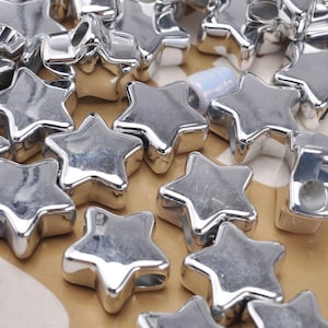 May include: A collection of small, five-pointed star-shaped beads with a reflective, metallic silver finish. Each bead features a smooth, rounded surface and a central hole for threading, arranged in a scattered pile on a flat surface.