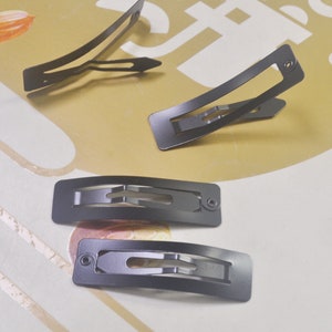 Könnte beinhalten: Vier schwarze Metall-Haarklammern. Die Klammern sind rechteckig mit einer gebogenen Form und haben ein kleines Loch oben.