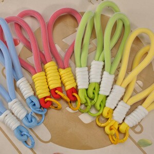May include: A collection of colorful keychains in various shades, including blue, pink, green, and yellow. Each keychain features a looped rope design with a contrasting color at the base and a metal clasp. The keychains are arranged on a patterned surface.