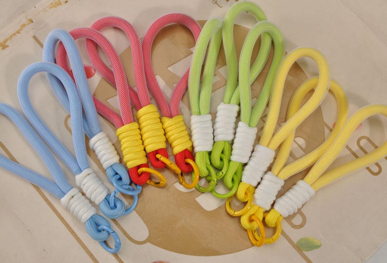 May include: A collection of colorful keychains in various shades, including blue, pink, green, and yellow. Each keychain features a looped rope design with a contrasting color at the base and a metal clasp. The keychains are arranged on a patterned surface.