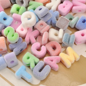 Puede incluir: Una colección de letras del alfabeto coloridas y difusas en varios tonos pastel, incluyendo rosa, azul, verde y amarillo. Las letras son suaves y afelpadas, con una apariencia texturizada. Las letras están esparcidas sobre una superficie marrón claro.