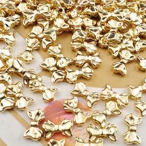May include: A large quantity of gold-colored metal bow charms. The charms are small and delicate, with a slightly raised design. They are perfect for adding a touch of elegance to jewelry, crafts, and other projects.
