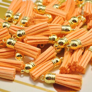 Könnte beinhalten: Eine Sammlung von orangefarbenen Wildlederquasten mit goldfarbenen Metallkappen und Schlaufen. Die Quasten sind in einem verstreuten Muster angeordnet, das ihre weiche Textur und lebendige Farbe zeigt. Diese kleinen dekorativen Elemente sind ideal für die Schmuckherstellung und Bastelprojekte.