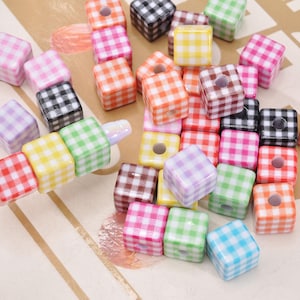 May include: A collection of colorful, square beads with a gingham pattern in various colors, including pink, green, yellow, and black. Some beads are strung on a metal rod. These beads are suitable for jewelry making and crafts.