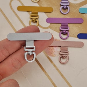 K&ouml;nnte beinhalten: Nahaufnahme eines silberfarbenen Handyhalters aus Metall, mit rechteckiger Oberseite und D-Ring. Weitere farbige Handyhalter in Gold, Lila und Ros&eacute;gold sind im Hintergrund zu sehen.