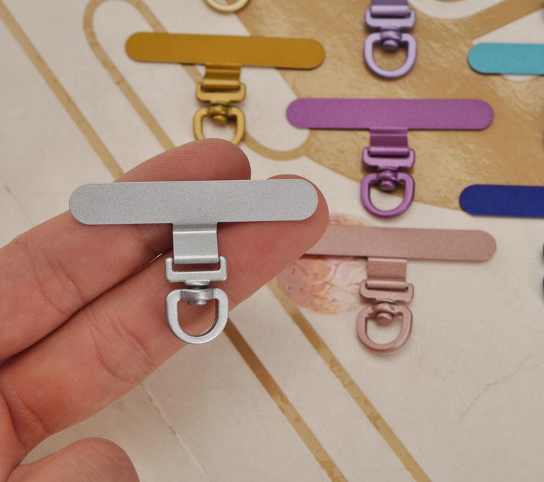 K&ouml;nnte beinhalten: Nahaufnahme eines silberfarbenen Handyhalters aus Metall, mit rechteckiger Oberseite und D-Ring. Weitere farbige Handyhalter in Gold, Lila und Ros&eacute;gold sind im Hintergrund zu sehen.