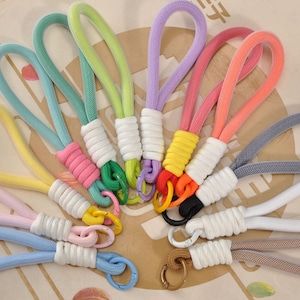 May include: A collection of colorful wrist keychains. Each keychain features a loop of woven rope in various colors, including blue, pink, yellow, and green. The rope is attached to a white wrapped base and a metal clasp.