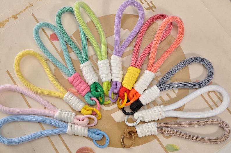 May include: A collection of colorful wrist keychains. Each keychain features a loop of woven rope in various colors, including blue, pink, yellow, and green. The rope is attached to a white wrapped base and a metal clasp.